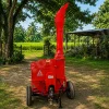 Red portable wood chipper on wheels in a rural yard, with a tall discharge chute and green fields in the background.