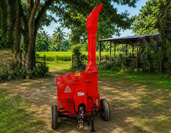 Red portable wood chipper on wheels in a rural yard, with a tall discharge chute and green fields in the background.